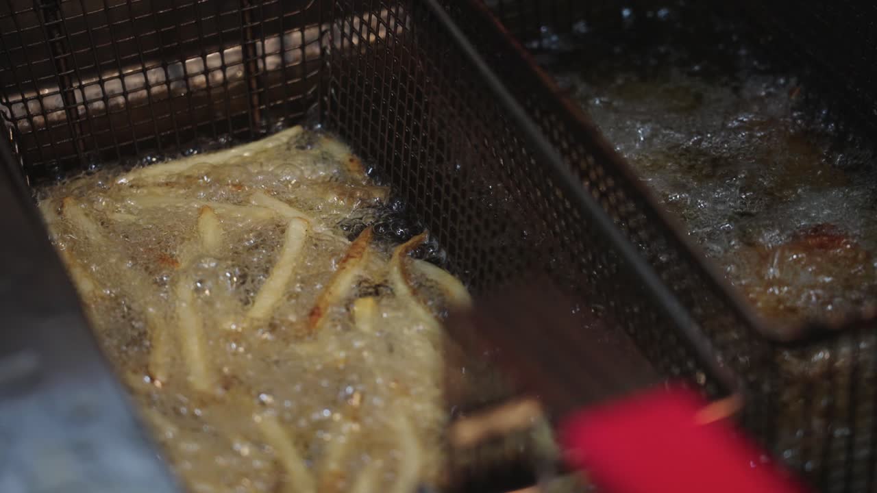 4K parallakseoptagelser af pommes frites og hot wings, der frituresteges i boblende varmt fedt, som fanger sydende olie, sprød tekstur og aktiv madlavning i et storkøkken.