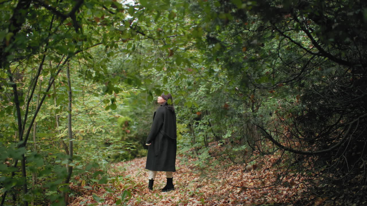 나뭇잎이 무성한 길에서 가을 나무 사이를 지나며 위를 올려다보고 나뭇가지를 스치는 캐주얼한 젊은 여성을 확대하여 보여 줍니다. 긴 코트가 흔들리고, 조용한 숲 터널을 탐험하는 평온한 방랑자, 황금빛 나뭇잎, 부드러운 빛이 있네요.