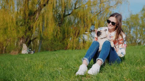 young woman resting in the park, sitting on a green lawn. he keeps a pug puppy on his lap, plays with a dog. has a good time