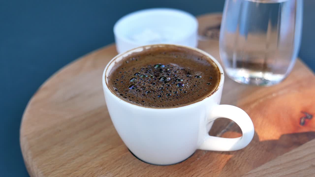 крупный план чашки турецкого кофе с пеной сверху, на деревянном подносе с стаканом воды и сахара