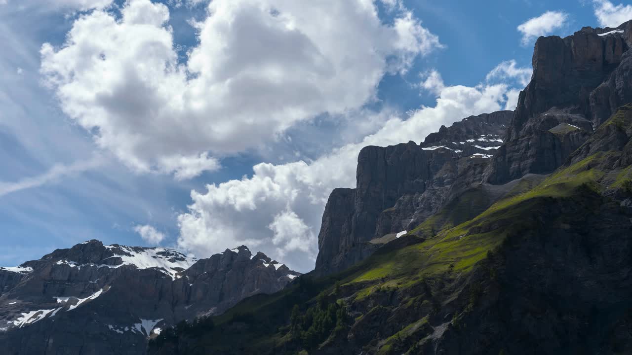 Time-lapse in Valais. mountains are partly covered in snow and grass. Clouds drift across the mountain range and swirl at the peak. Shadows cover the rock. The sky is blue. Epic shot in the Swiss Alps