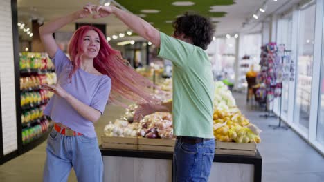 Happy-brunette-guy-with-his-girlfriend-with-pink-hair-dancing-and-holding-hands-in-a-grocery-store