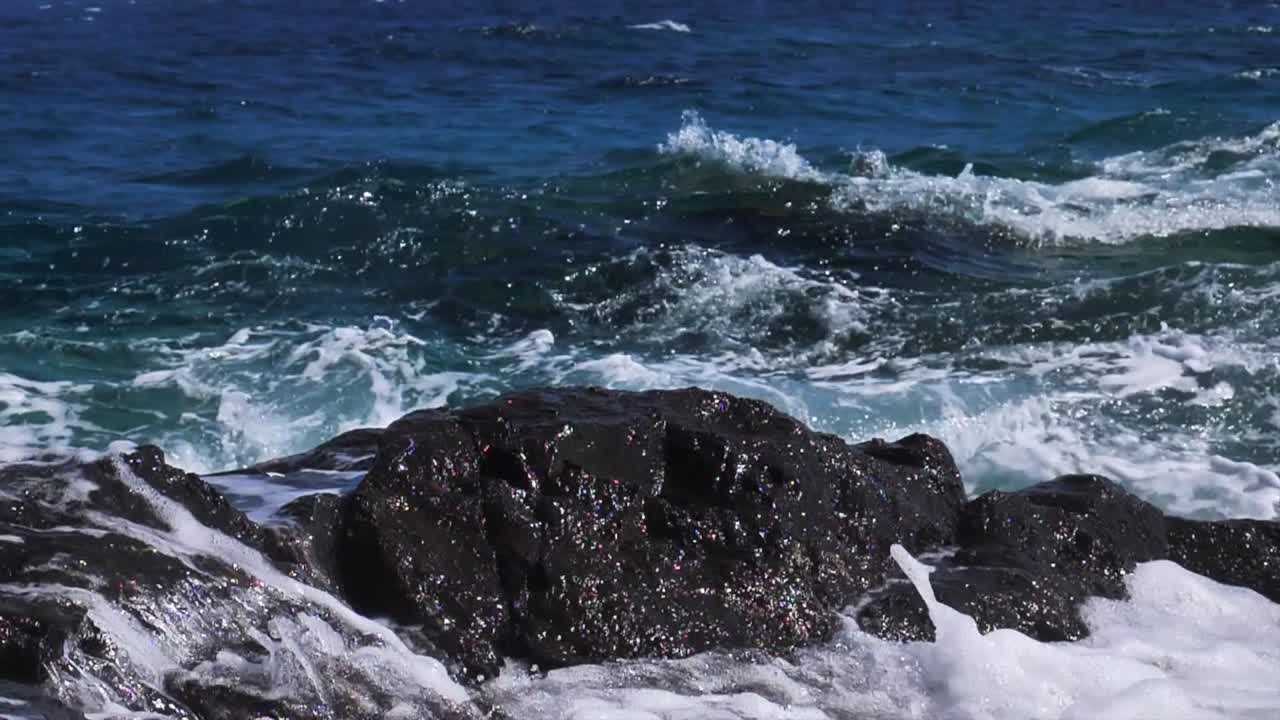 Ripresa al rallentatore di onde oceaniche che si infrangono contro scogliere costiere scure in Cornovaglia, Inghilterra, mostrando schiuma bianca, il movimento dell'acqua marina e la texture frastagliata della costa.