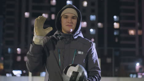 man in hoodie and gloves casually hitting a soccer ball in his hand while standing on an outdoor urban sports field, nighttime scene illuminated by bokeh lights and surrounding high-rise buildings