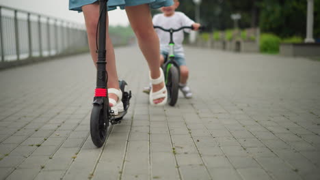 a woman rides a black scooter on a paved walkway, her legs visible as she moves ahead, a child follows closely behind her on a small bicycle