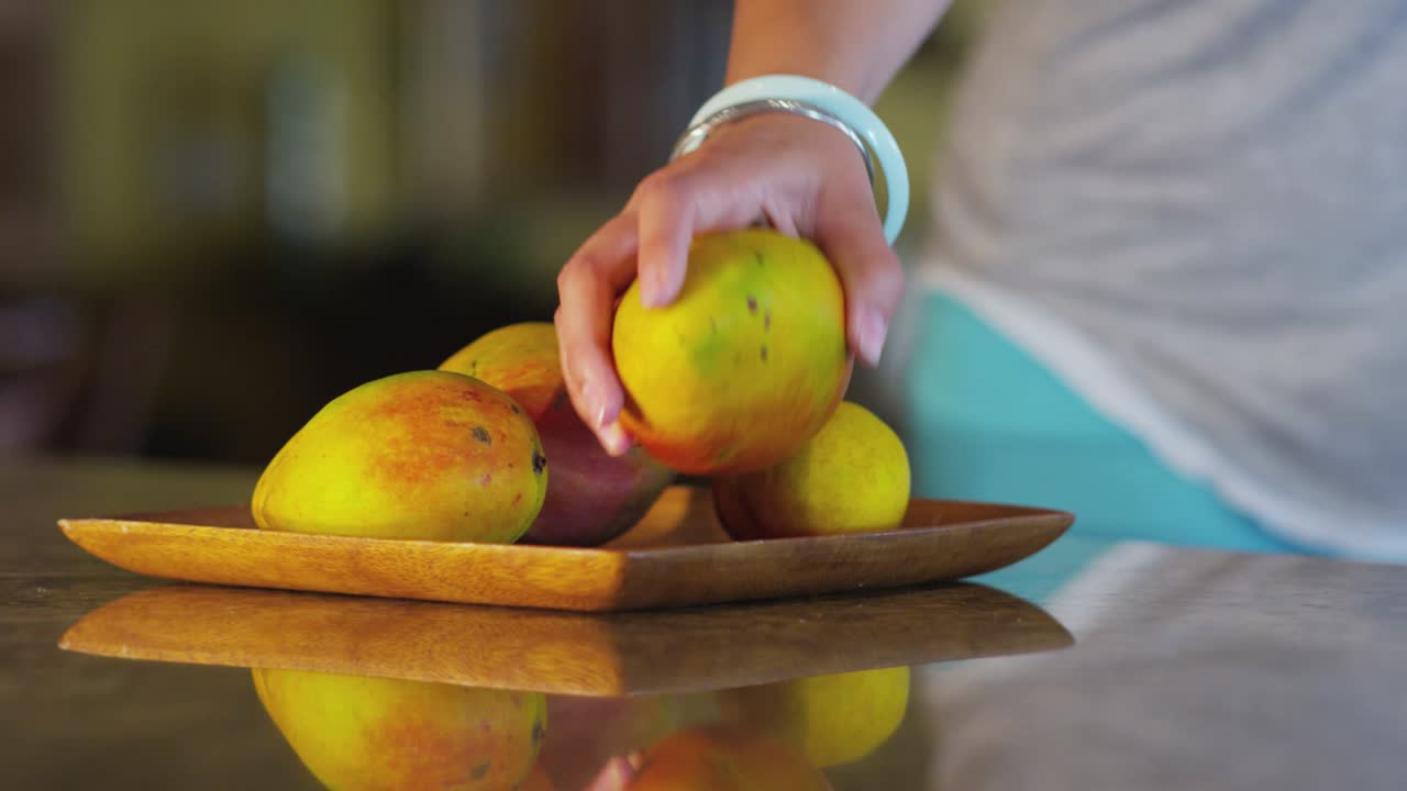 Zbliżenie przedstawiające rękę osoby wybierającej dojrzałe mango z drewnianego talerza wypełnionego świeżymi owocami. Talerz spoczywa na odbijającej światło powierzchni, odbijając żywe kolory owoców i ruch ręki.