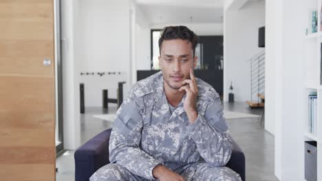 serious biracial male soldier in uniform sitting in armchair at home having video call, slow motion