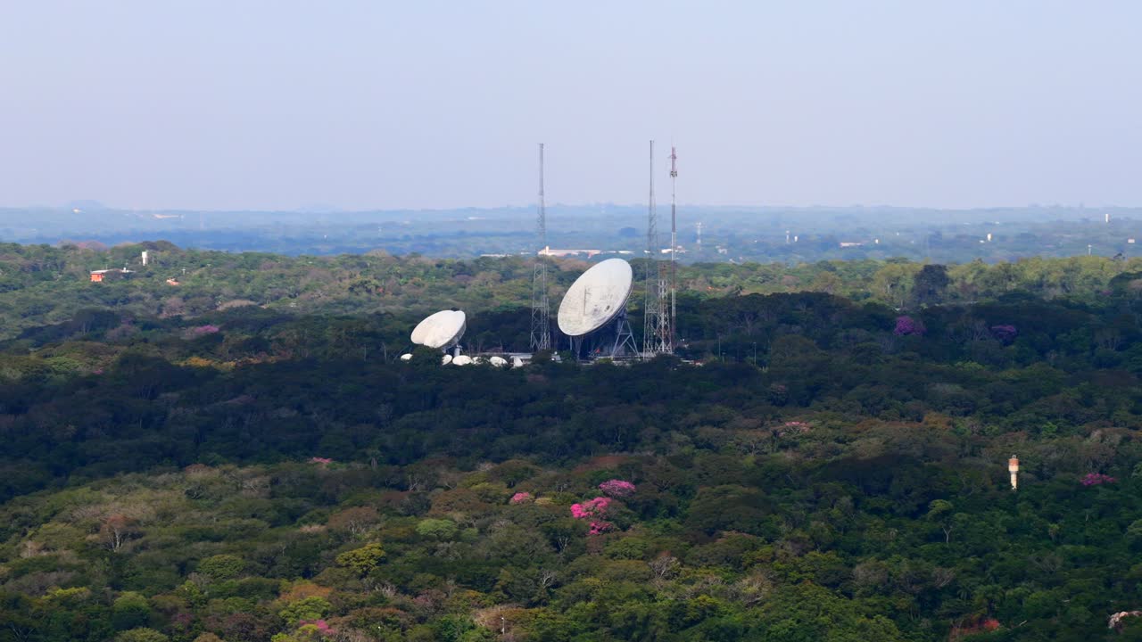 Drone footage captures expansive view of satellite dishes nestled in lush Areguá forest. The serene setting features vibrant green foliage, clear skies, and advanced technology