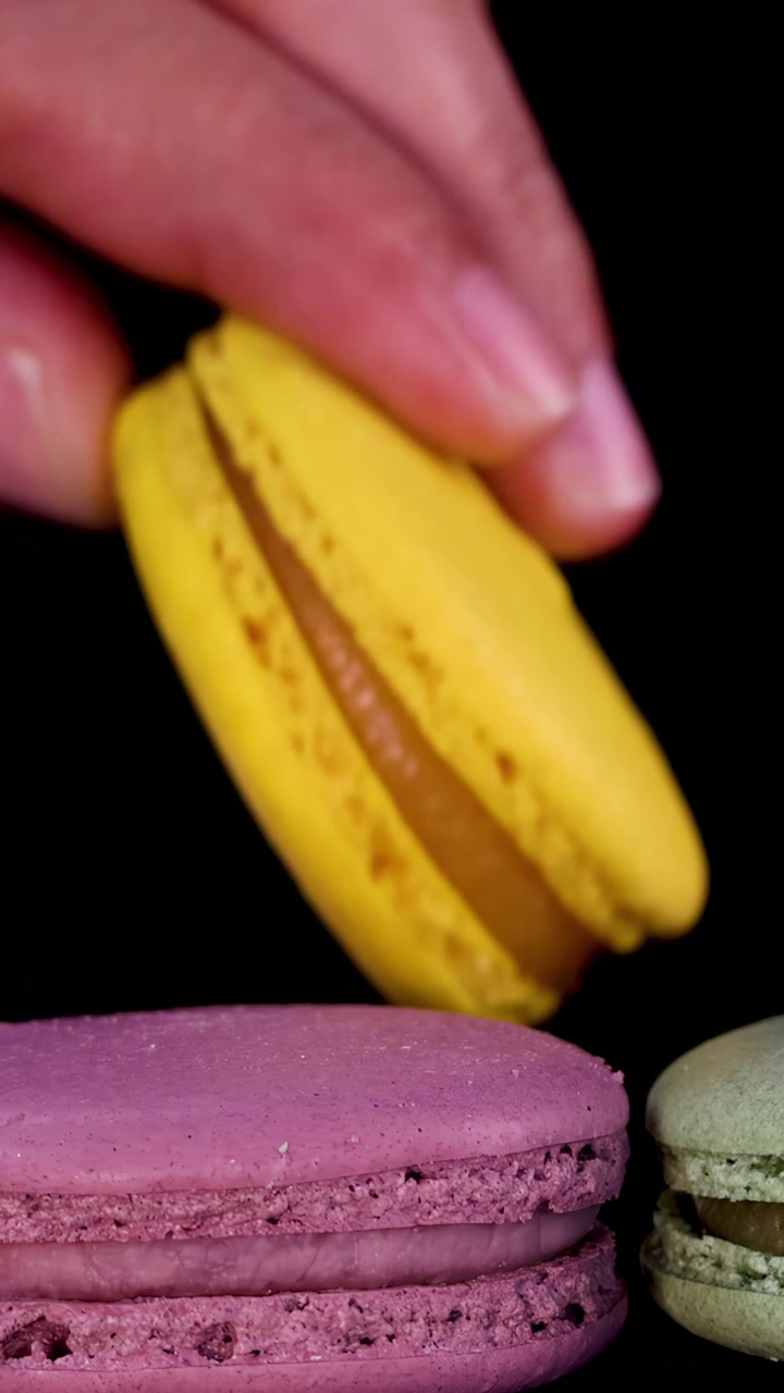 A hand reaches in to pick up a yellow macaron from a neat row of assorted colorful macarons on a black background, under bright studio lighting with a close-up perspective