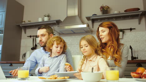 Cheerful-Family-Having-Breakfast-While-They-Use-A-Laptop-And-A-Tablet