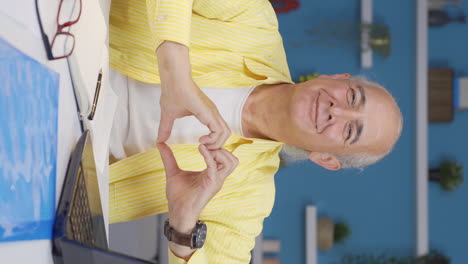 Vertical-video-of-Home-office-worker-old-man-makes-heart-symbol-looking-at-camera.