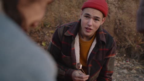 camera focuses on a teenage boy roasting marshmallows over the fire while talking to a friend who is sitting in front of him