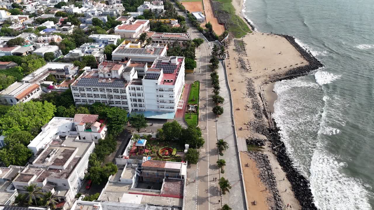 Aerial view of Puducherry, a well-developed coastal city with a modern aesthetic. The sea borders a wide, sandy beach. there is a mix of large, modern buildings, with solar panels on their rooftops