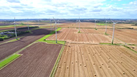 A-Vista-De-Pájaro,-Observamos-Las-Turbinas-Eólicas-Girando-Elegantemente-En-El-Campo-Recién-Cosechado-De-Un-Granjero-De-Lincolnshire,-Complementadas-Con-La-Presencia-De-Fardos-De-Heno-Dorados.