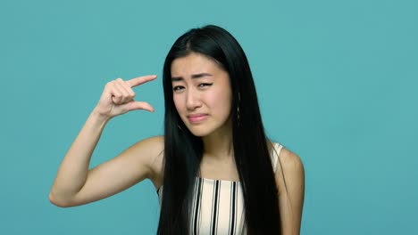 positive asian woman with long black hair making little bit gesture, measuring small size and looking at camera