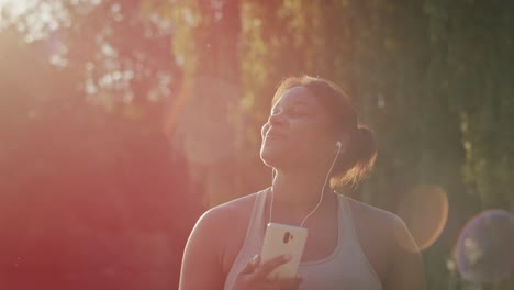 Plus-sized-African-American-woman-walking-and-using-mobile-phone-with-earphones-in-the-park-in-a-summer-day