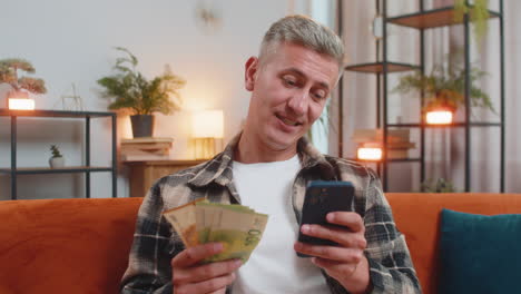 smiling happy caucasian man counting money euro cash use smartphone income salary lottery game win
