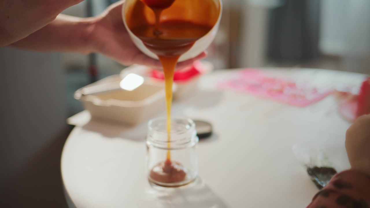 Mani versano sciroppo dorato in un barattolo di vetro su un tavolo bianco. Una luce calda evidenzia il flusso del ricco liquido ambrato. La scena mostra un'azione attenta e deliberata. Perfetto per progetti di cibo, bevande o artigianato.