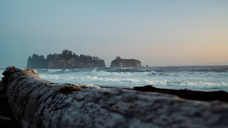 Gorgeous-landscape-beach-shot-of-driftwood-with-waves-crashing-in-the-background,-a-beautiful-small-cliff-island,-and-a-colorful-cloudy-sky-during-sunset-at-the-famous-Ruby-Beach-in-Forks,-Washington