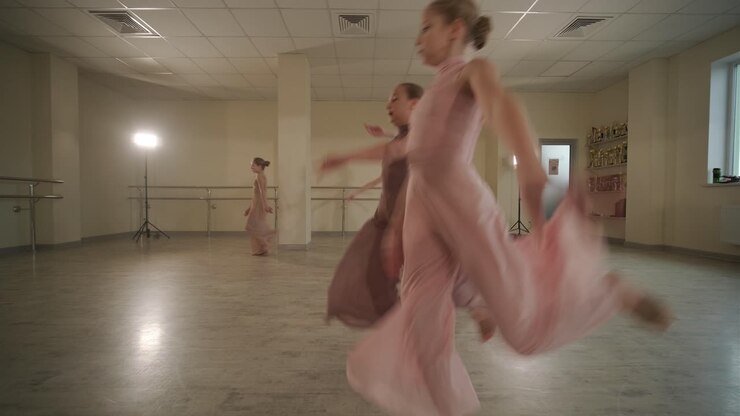 Un gruppo di giovani allievi di danza classica in abbigliamento da ballo nero che eseguono esercizi in un ampio studio di danza con pavimenti in legno e barre a muro. Espressioni concentrate e movimenti sincronizzati