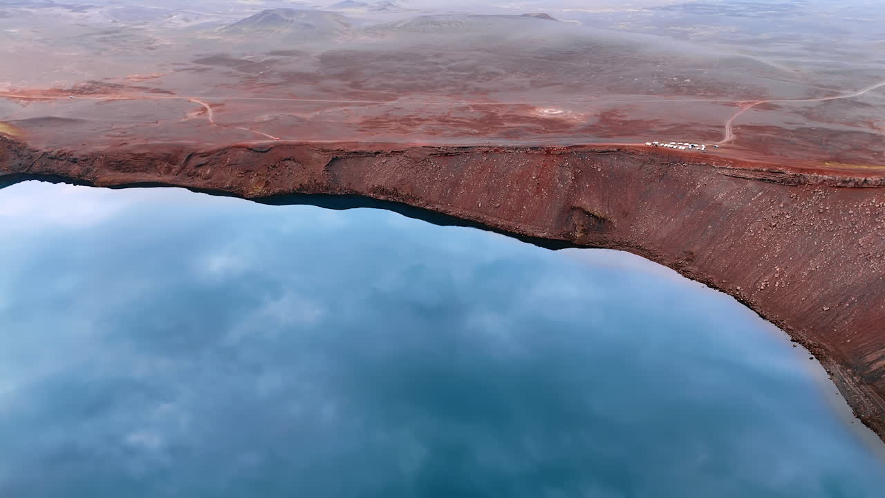 Blue sky reflects in the lake located in the sleeping crater volcano. Drone footage above the rocky landscape of Iceland.