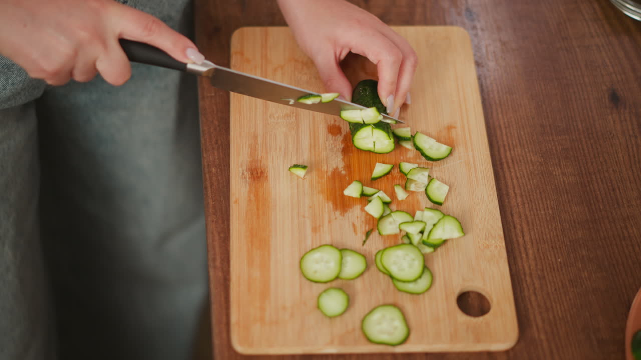 Overhead view of white girl slicing cucumber on wooden board with glass bowl of cucumber slices nearby on countertop while preparing healthy meal ingredients in bright modern kitchen setting