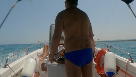 back view of man with blue swimwear driving fast motorboat steering wheel with people sunbathing on bow