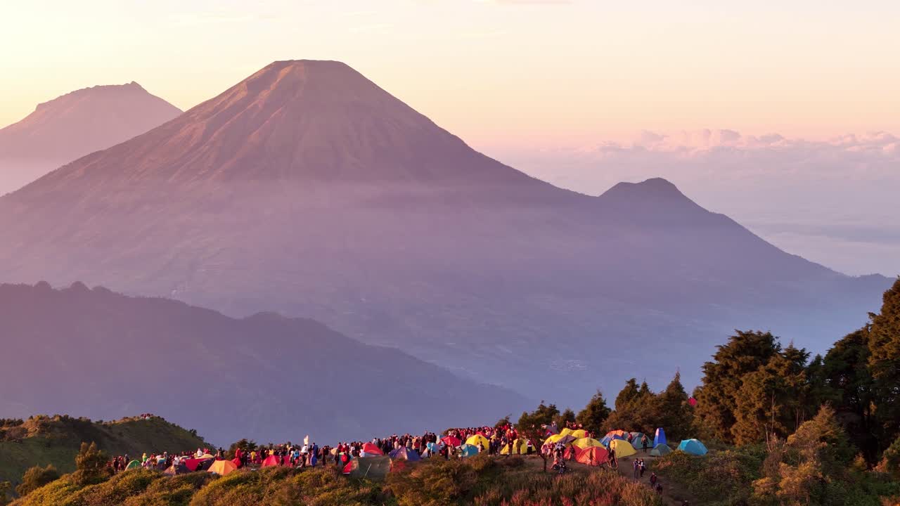 Vista aerea di un campeggio colorato in cima a una montagna, brulicante di escursionisti all'alba, con uno spettacolare paesaggio montano e strati di colline nebbiose sullo sfondo. Monte Prau, Indonesia