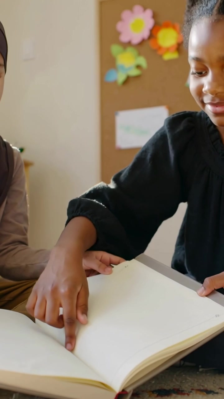 Vertical video: Reaching right child turning pages at school, sharing blank book, left in headscarf