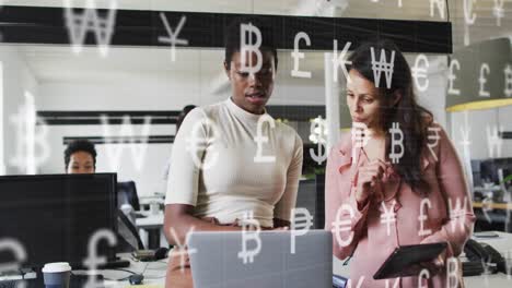 Animation-of-currency-symbols-against-two-diverse-women-discussing-at-office