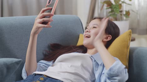 close up of asian teen girl having video call on smartphone while lying on sofa in the living room at home. waving hands, smiling, and speaking