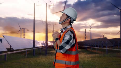 side view of asian male engineer with safety helmet pray for something while standing with solar panel and wind turbines