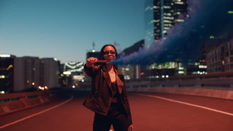 young woman holding blue smoke bomb in city at night