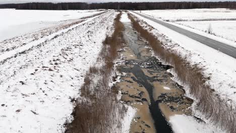 Drohne-Folgt-Genau-Dem-Winterkanal-Mit-Halb-Gefrorenem-Schmutzwasser-Und-Verschneiter-Landschaft