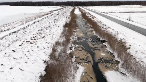 drone closely follows winter canal with half frozen dirty water, snowy landscape