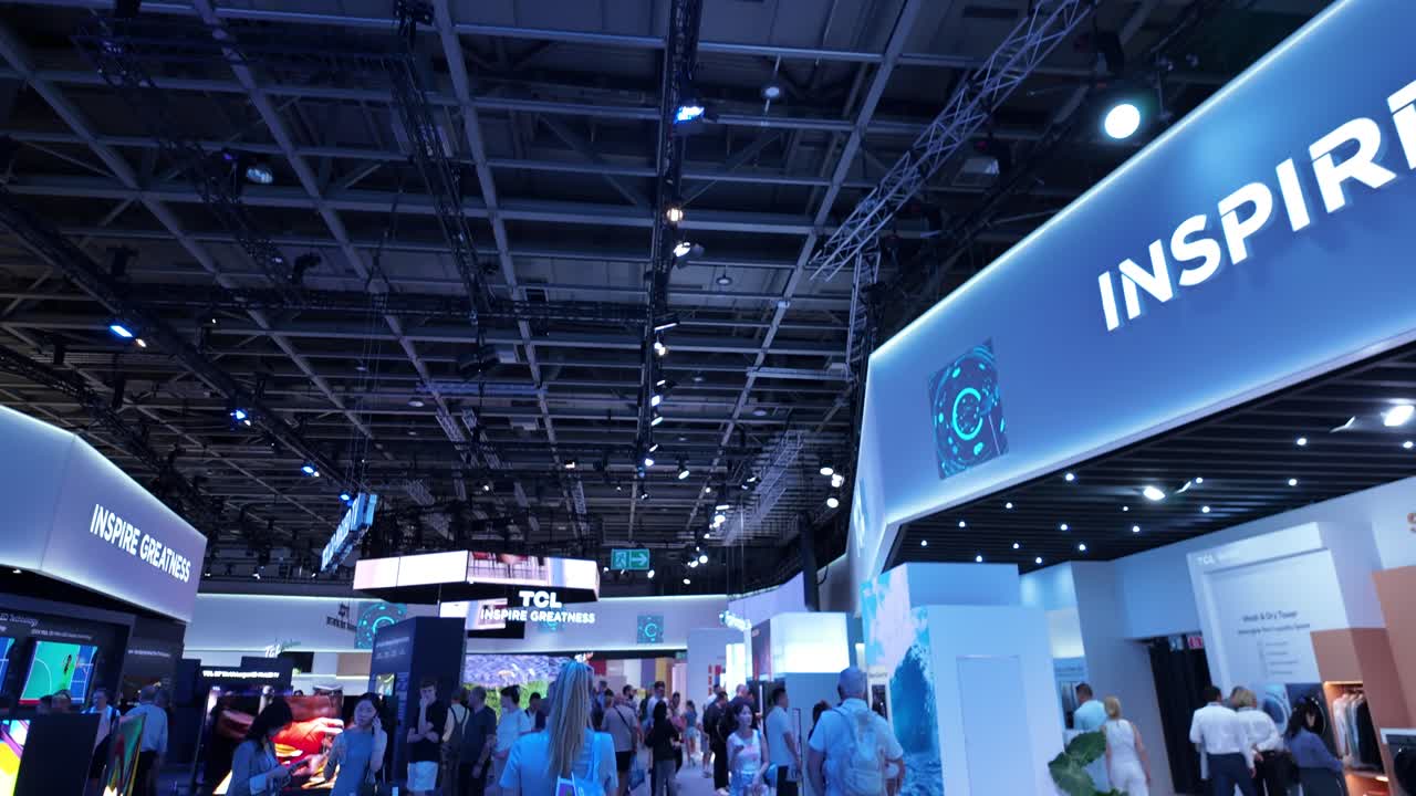 IFA Berlin tech expo bustling with attendees exploring futuristic displays and innovations from leading brands. The blue-lit ambiance enhances the tech-focused environment