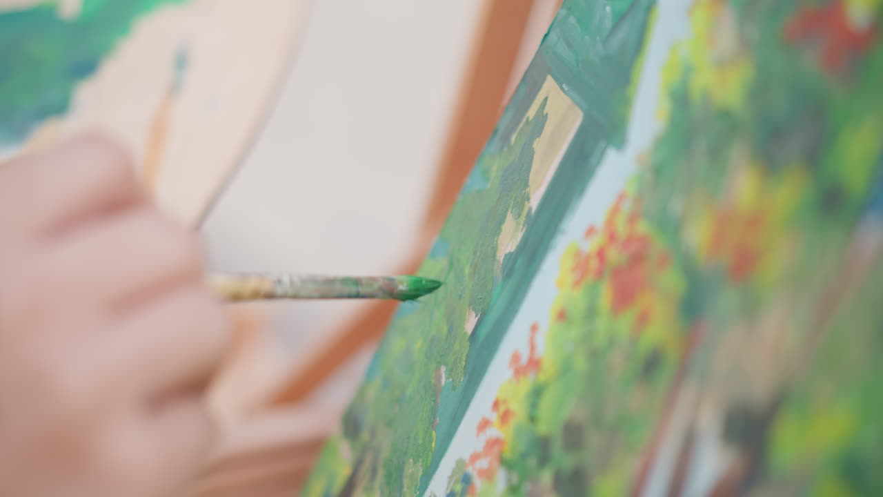 primo piano di un dipinto a pennello di un vivace paesaggio verde su area di lavoro. Una mano tiene un pennello rotondo applicando pennellate materiche a un paesaggio floreale su un cavalletto; una palette si intravede sullo sfondo, un artista di strada che perfeziona uno schizzo.