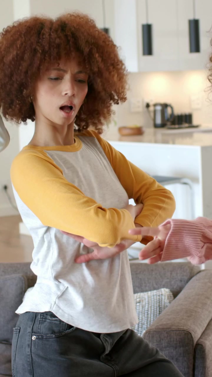 Vertical video: Shifting weight, curly woman dancing at home by sofa for friend, wearing raglan