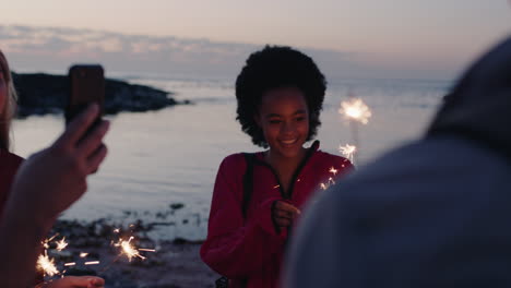 jovem loira tirando fotos de amigos segurando fogos de artifício comemorando a véspera de ano novo usando smartphone aproveitando a festa na praia ao pôr do sol