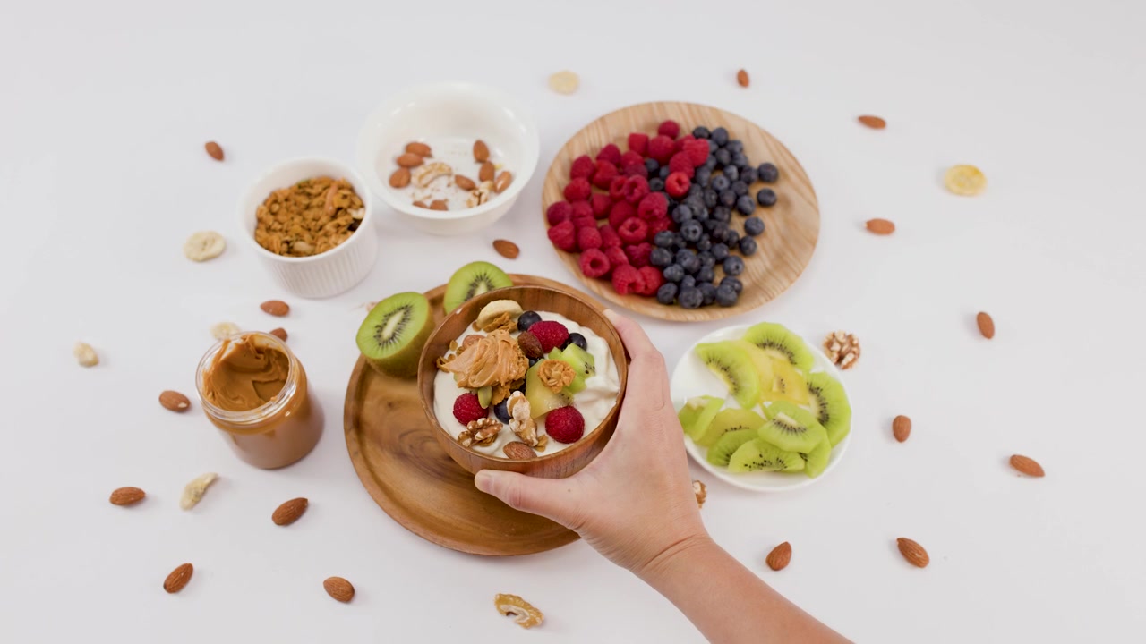 Tangan meletakkan mangkuk yogurt kayu dengan buah dan kacang di atas permukaan putih dengan bahan-bahan