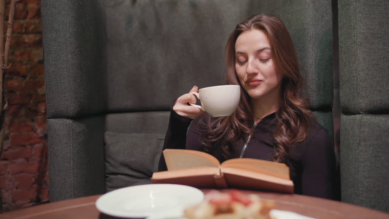 Lady in black sits relaxed inside booth with brick wall background, reading brown book and flipping page while sipping drink from white cup, surrounded by cozy ambiance and waffle on plate