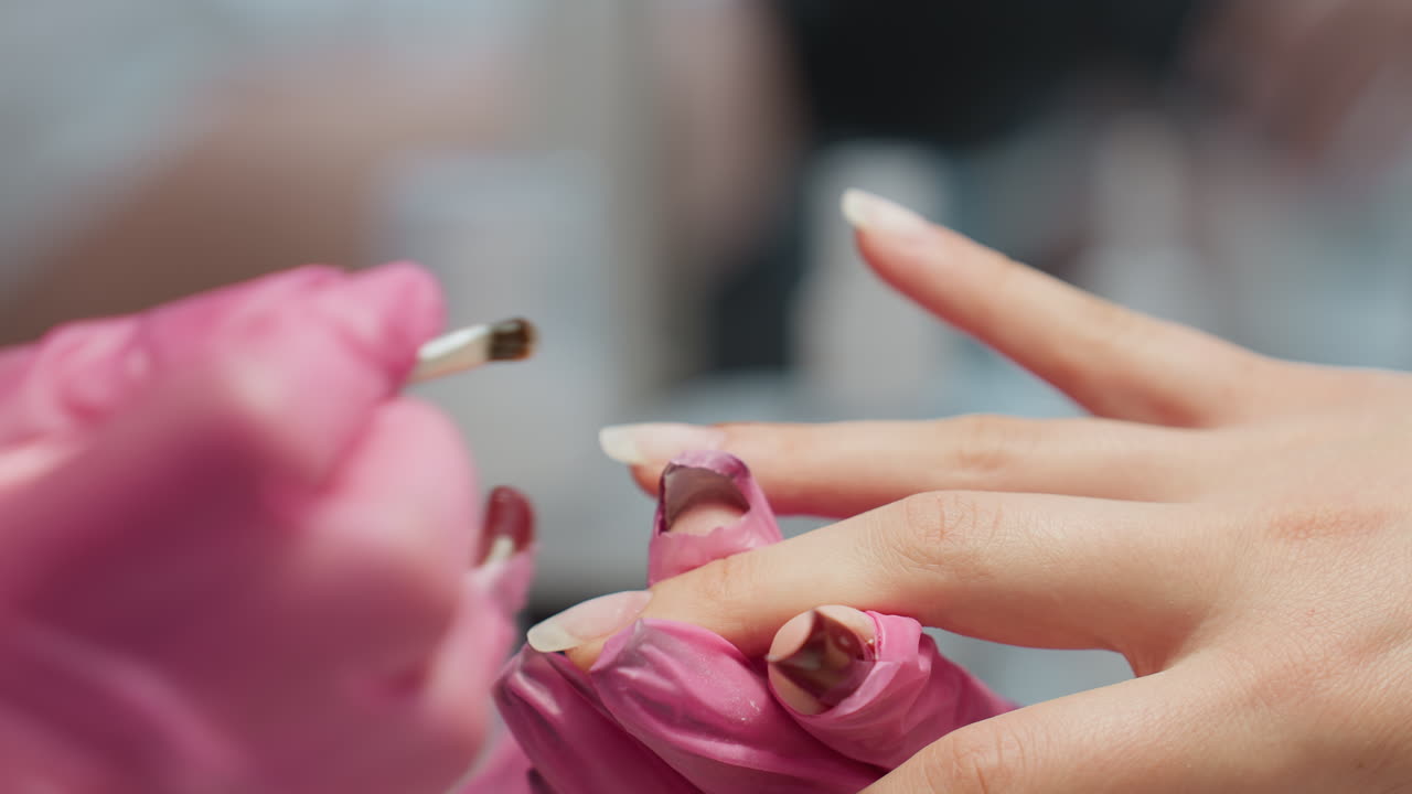 Close up view of nail technician wearing pink gloves carefully applying hardener with brush to client fingernail during manicure session, with soft blur background emphasizing detail and precision