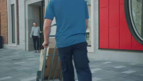 delivery man pushes hand truck trolley full of cardboard boxes hands package to a customer, who then signs electronic pod device. courier delivers parcel to man in business district. following shot