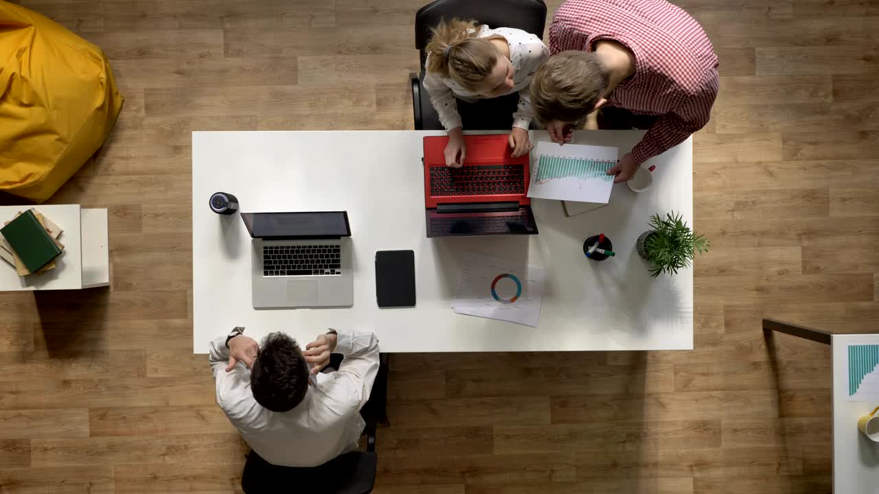 Mand diskuterer projekt med kvinde, mens en anden mand arbejder på bærbar computer og taler i telefon, topskud, moderne kontorbaggrund