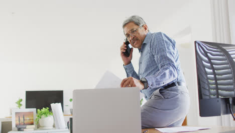 Video-of-senior-biracial-man-sitting-on-desk-talking-on-phone-and-using-laptop-at-home