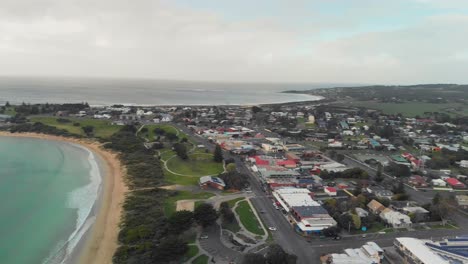 -panning-shot-of-the-town-of-Apollo-Bay-showing-the-layout-of-the-streets-and-buildings-situated-on-a-beautiful-stretch-of-coastline,-surrounded-by-lush-green-forests-and-rolling-hills