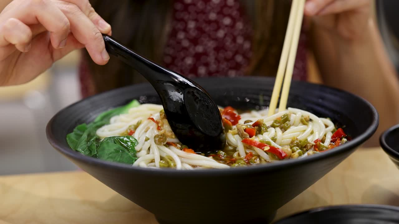 Woman stirs hot spicy noodle soup with chopsticks and spoon in bright restaurant setting