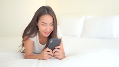 young smiling beautiful asian woman lying on bed chats on smartphone