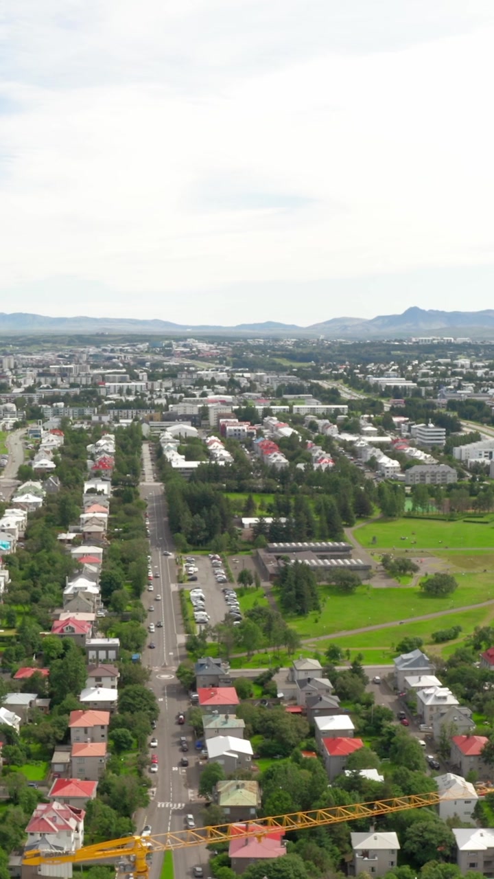Una ripresa aerea con drone sorvola un quartiere residenziale a Reykjavik, Islanda, mostrando case colorate, una gru da costruzione e montagne distanti sotto un cielo luminoso