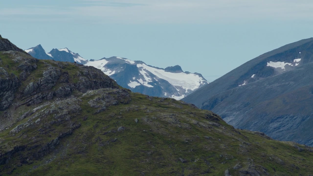 Lagdelte steinete åser med grønne skråninger foran isbredekte fjelltopper under en blekblå himmel